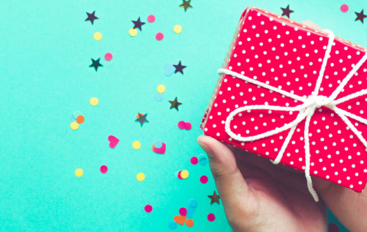 a small gift, wrapped in red paper tied with string, is held over a turquoise background with heart, star and circle shaped confetti 