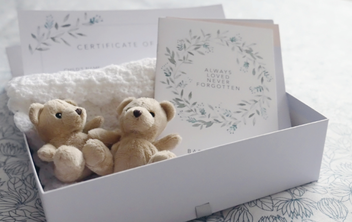 A white box with 2 small teddy bears placed on a white knitted blanket, and a card reading "always loved, never forgotten"