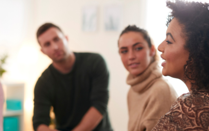 A woman with dark, shoulder length, curly hair is pictured from her left side talking to a group of people. A man and woman listen to her.
