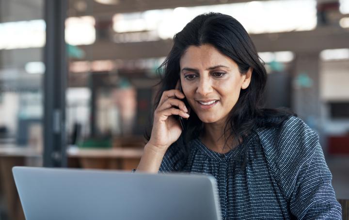 A lady sat at her desk, looking at her laptop whilst on the phone