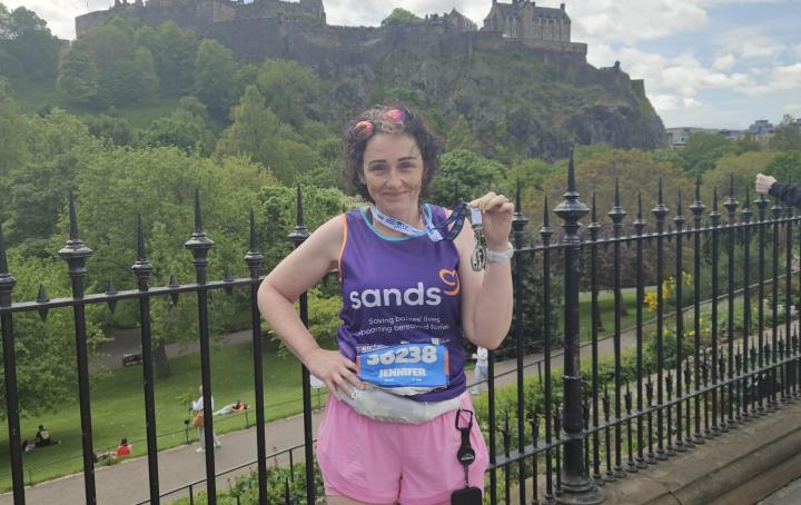 A woman standing holding a medal up with her hand, and Edinburgh Castle behind her.