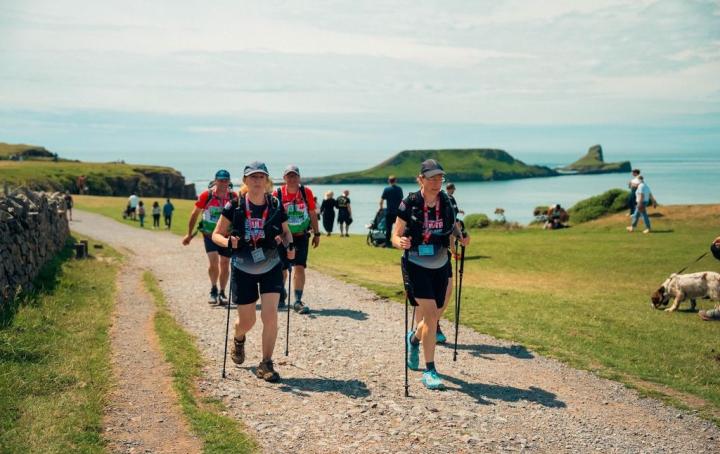 A group of people walking on a gravel path, in the background there is green grass, dog walkers and the irish sea.
