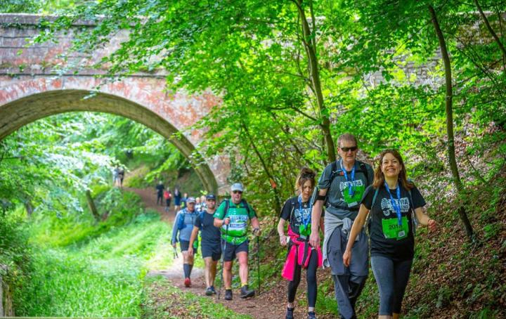 A long line of people walking on a path, in the background there are trees with green leaves on a hill and an orange brick bridge.