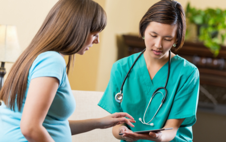 A woman wearing a light green t-shirt gestures toward a tablet device held by a female healthcare worker that they are both looking at 