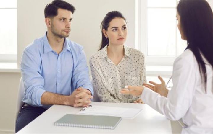 A white man withshort dark hair wearing a blue shirt and a white woman with long dark hair wearing a beige shirt sit at a desk facing a female healthcare worker with long dark hair wearing a white lab coat. She has her back to the camera and is gesturing with her hands. The man and woman look serious but calm.