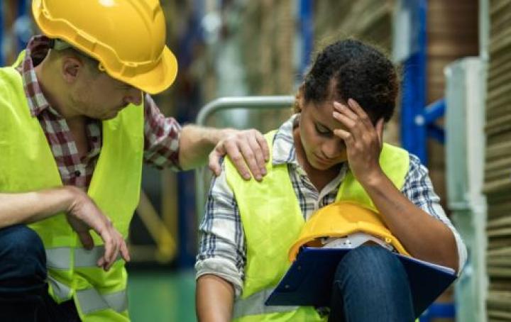 A white male warehouse worker in a red check shirt, hi-vis jacket and yellow hard hat puts their hand comfortingly on the shoulder of a Black female co-worker wearing a blue check shirt and hi -via jacket. She is sitting with one hand on her forehead and looks upset.