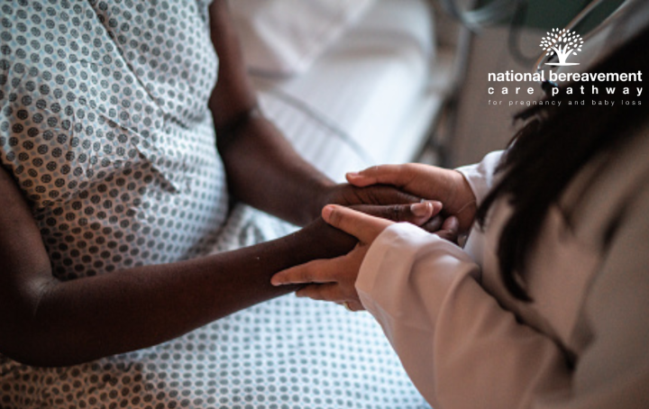 Black woman in hospital gown is sat on a bed, her hands held by a person with long hair and brown skin who is standing by her.