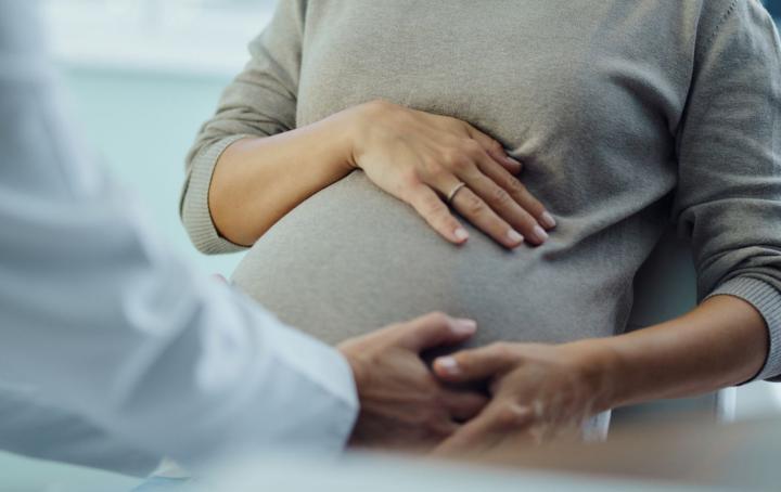 The arm of a health care worker in a white lab coat holds the left hand of a pregnant woman with her right hand on her stomach 