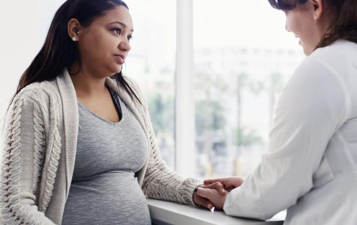 A pregnant woman wearing a white cardigan and grey t shirt is reassured by a female health worker in a white lab coat