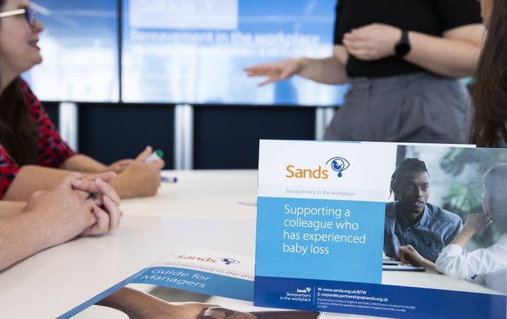 Supporting a colleague who has experienced baby loss flyer stood on the table with a lady sat down talking to someone who is stood in front of them in the background