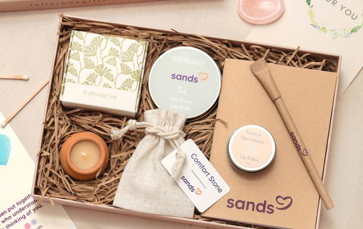 Sands Wellbeing Box pictured with notebook, pen, comfort stone, wildflower seeds, tea, tea light and lip balm