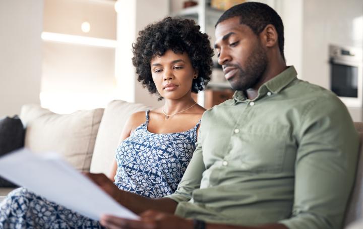 Black woman with afro and patterned blue top sits on sofa next to Black man with short hair and a green shirt, both looking at a piece of paper.