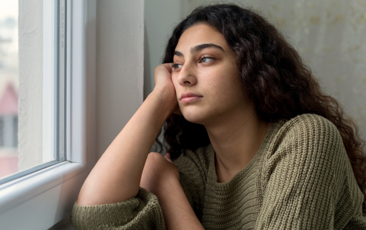 A young woman with long dark hair is looking out of a window with a sad expression