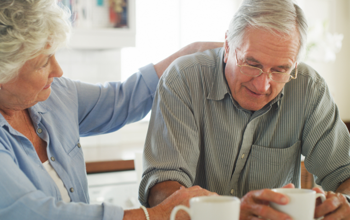 An older woman puts a comforting hand on the shoulder of an older man