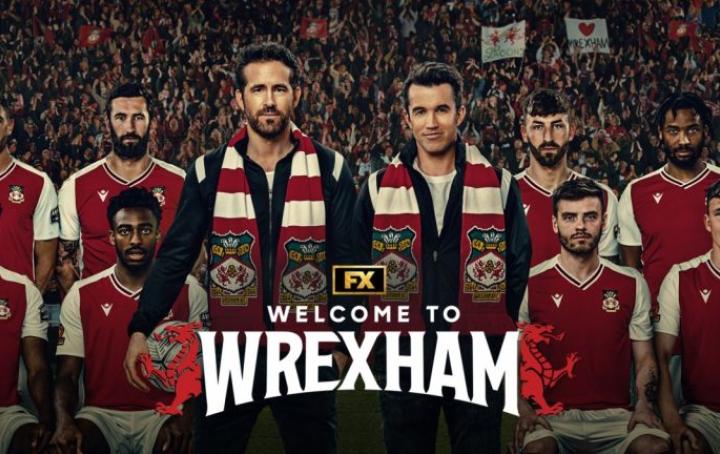 Show image of Rob McElhenny and Ryan Reynolds standing in front of Wrexham football team wearing club scarfs