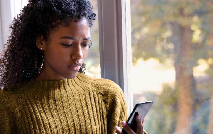 A Black woman wearing a yellow jumper looks at her mobile phone with an expression of concern