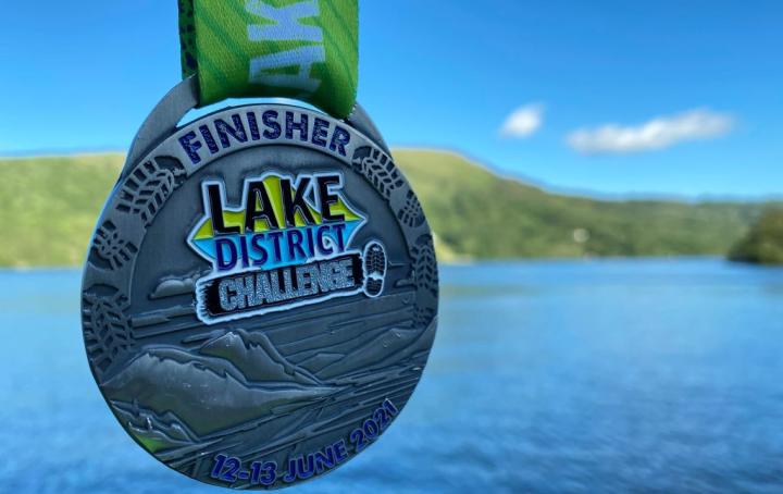 A medal with green ribbon, lsilver plate with the writing finisher medal Lake District Challenge, with Lake and green trees in the background.