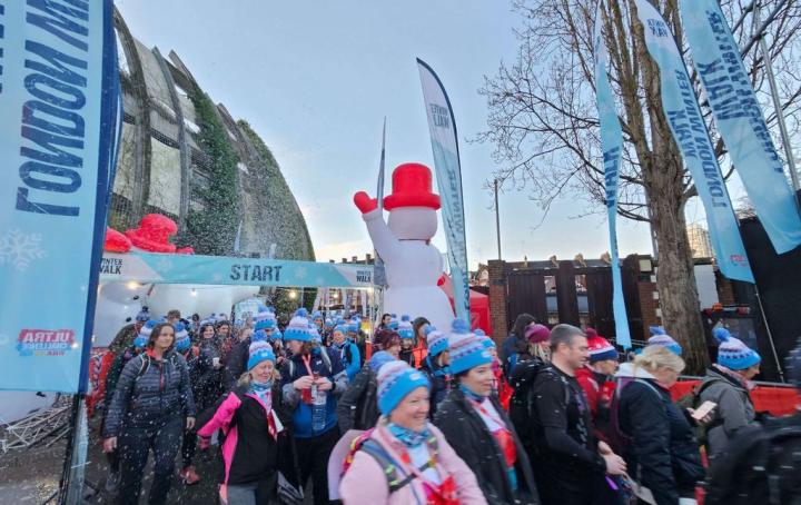 Large crowd of people walking, they are all wearing coats and blue bobble hats. There is a blow up snowman in the background.